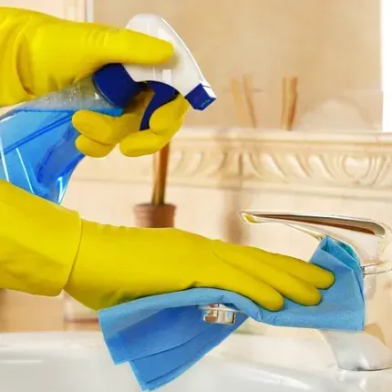 Persona que usa guantes amarillos rociando y limpiando un grifo del baño con un paño azul.