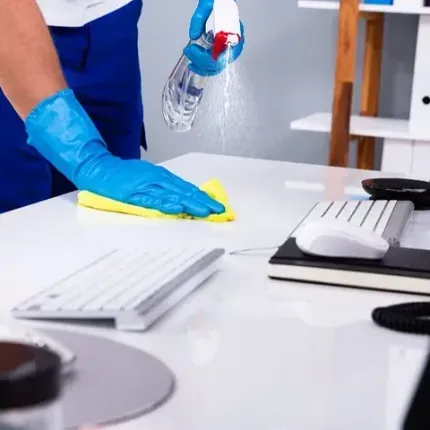 Persona con uniforme azul y guantes rocía y limpia un escritorio con teclado y ratón.