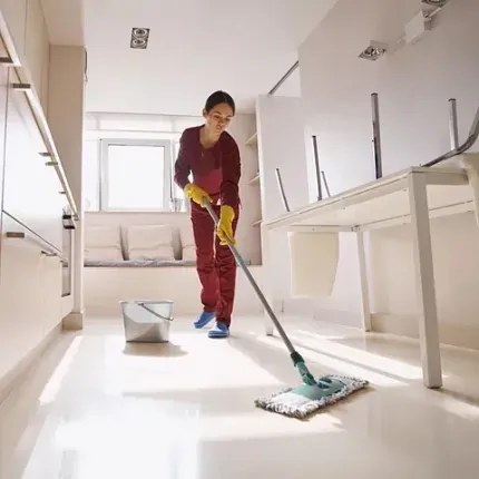 Mujer trapeando una habitación bien iluminada con gabinetes y paredes blancos; usa guantes amarillos y un traje color burdeos.