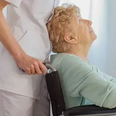 Un trabajador sanitario con uniforme blanco empuja a una mujer mayor en silla de ruedas; la mujer mira hacia arriba con una expresión amable.