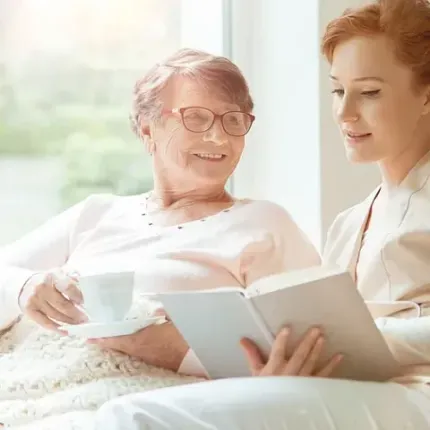 Una anciana y una joven leen un libro en un espacio cerrado junto a una ventana, sonriendo y mirándolo. La anciana sostiene una taza y un platillo.