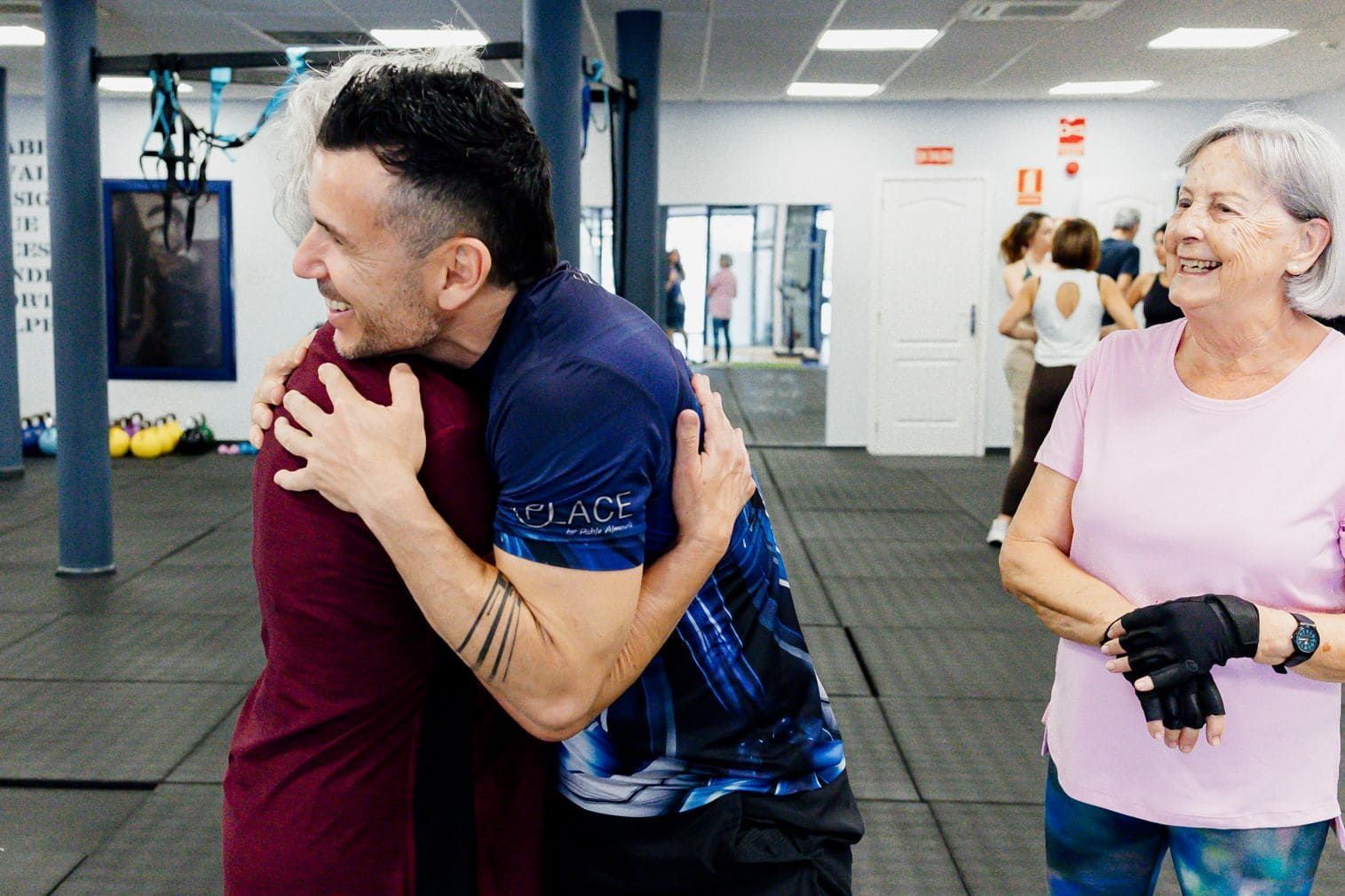 Un hombre abraza a una mujer mayor en un gimnasio, sonriendo. Otra mujer mayor observa con una sonrisa.