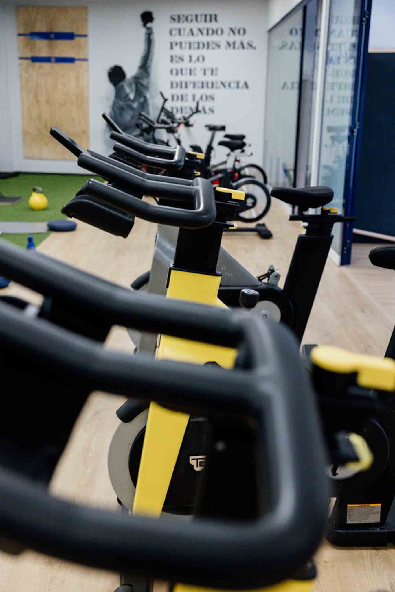 Bicicletas de spinning alineadas en un gimnasio con una cita motivacional en la pared y una pesa en el fondo.