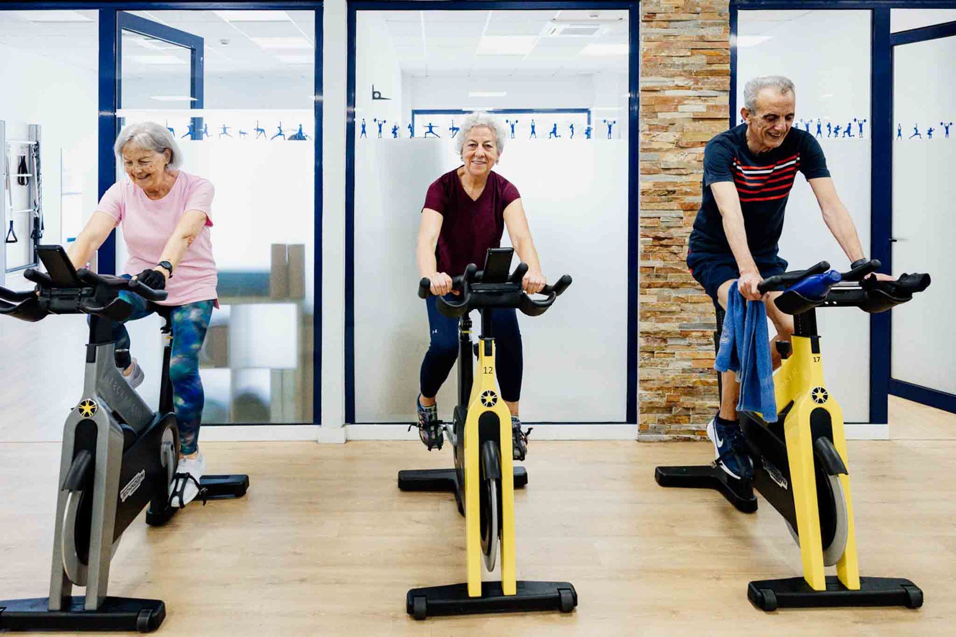 Tres adultos mayores sonríen mientras montan en bicicleta estática en un gimnasio. 