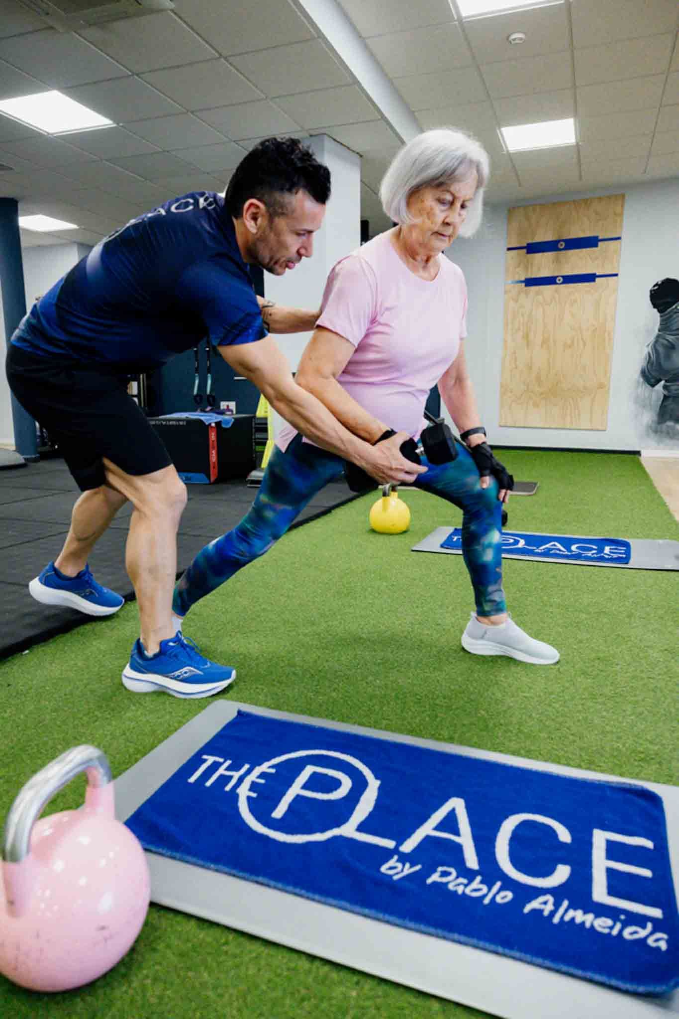 Un entrenador personal ayuda a una mujer mayor a hacer una zancada con una pesa rusa sobre el suelo verde del gimnasio.
