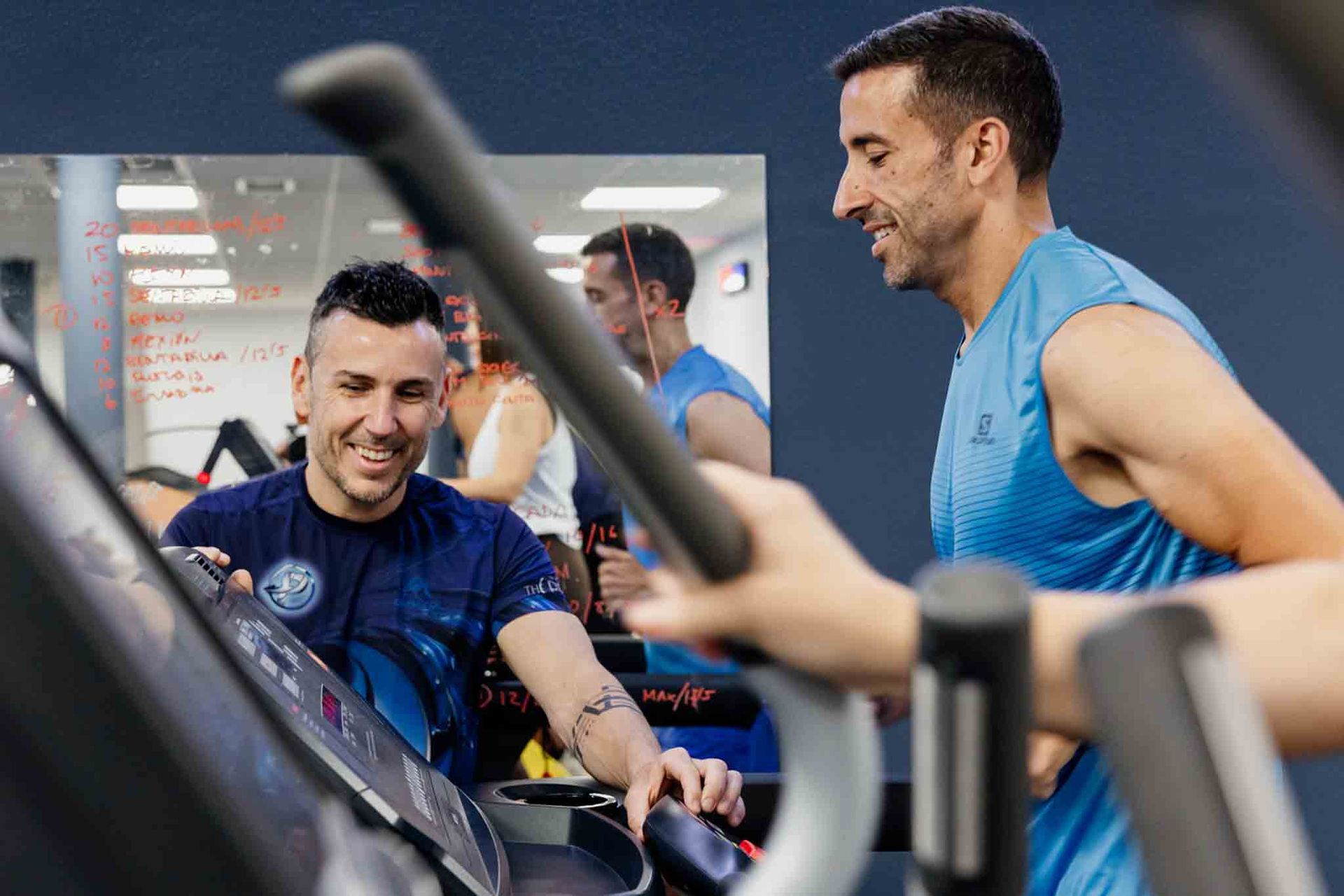 Dos hombres en un gimnasio; uno ajusta la consola de una cinta de correr mientras el otro corre, sonriendo. 