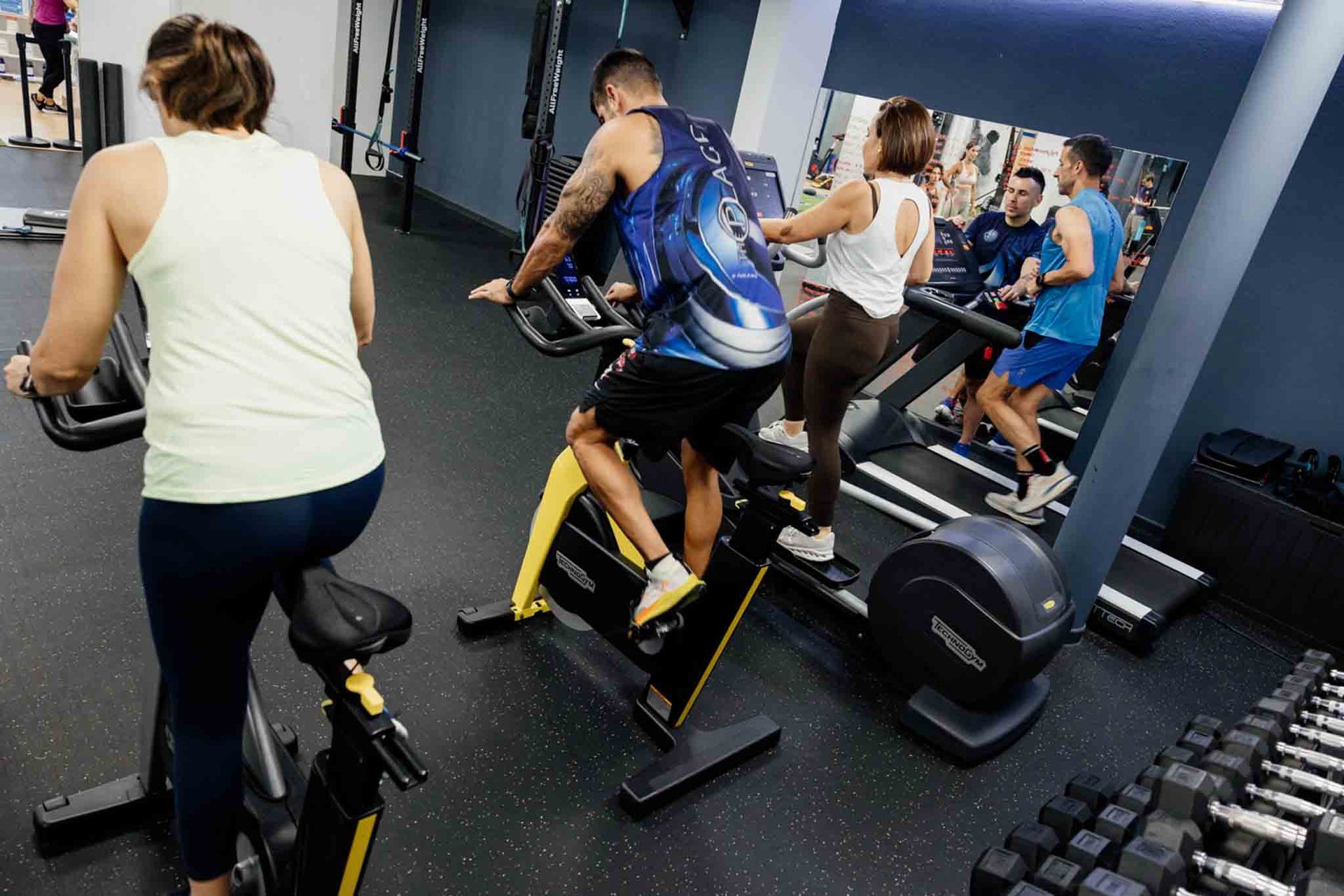 Personas haciendo ejercicio en un gimnasio: dos en bicicletas estáticas y dos en cintas de correr junto a un espejo. 