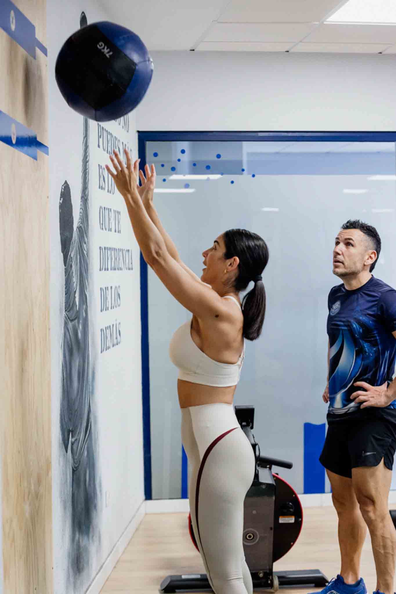 Una mujer vestida con ropa deportiva lanza un balón medicinal contra una pared mientras es supervisada.