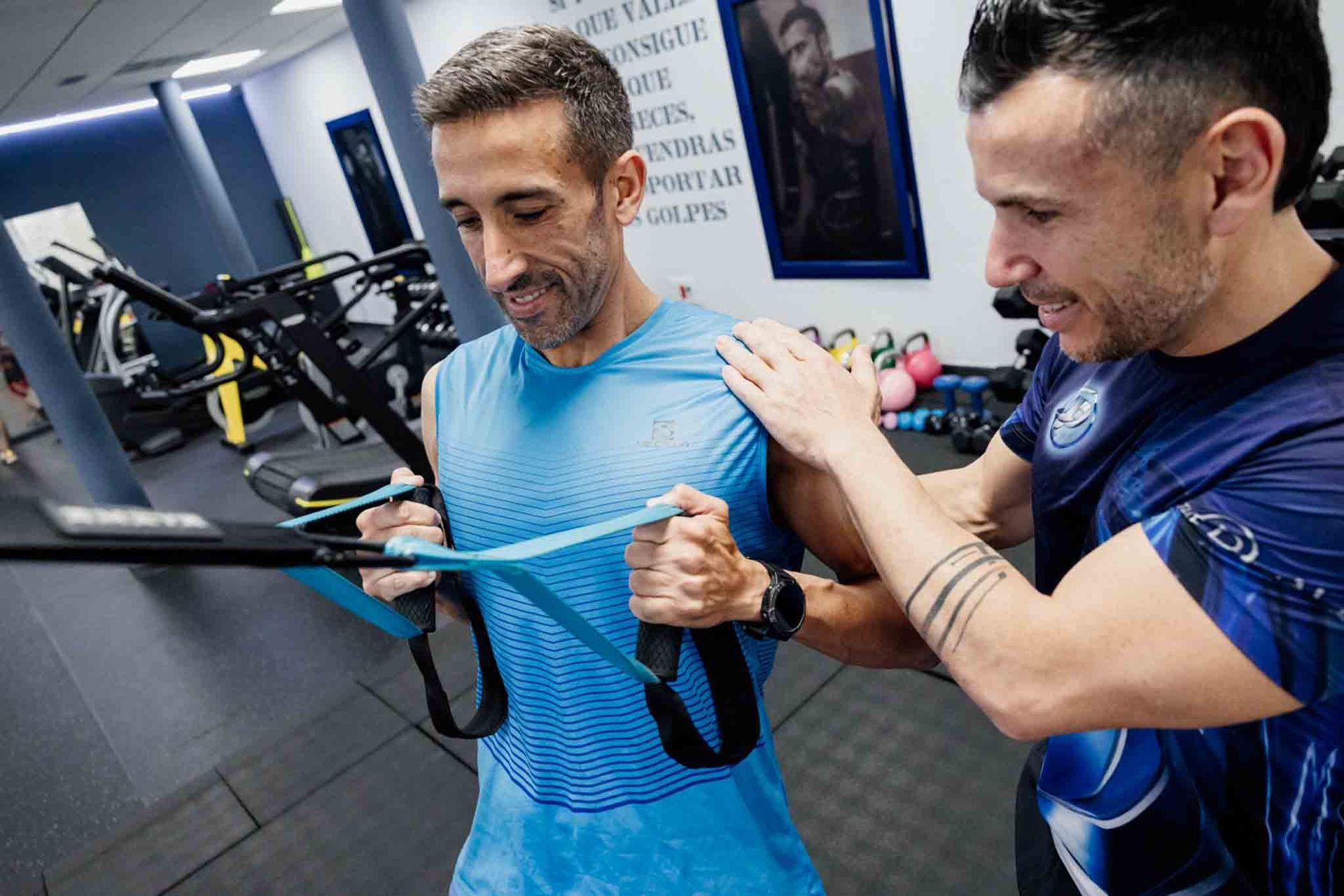 Un hombre con camisa azul se ejercita con una banda elástica, guiado por un entrenador en un gimnasio. 