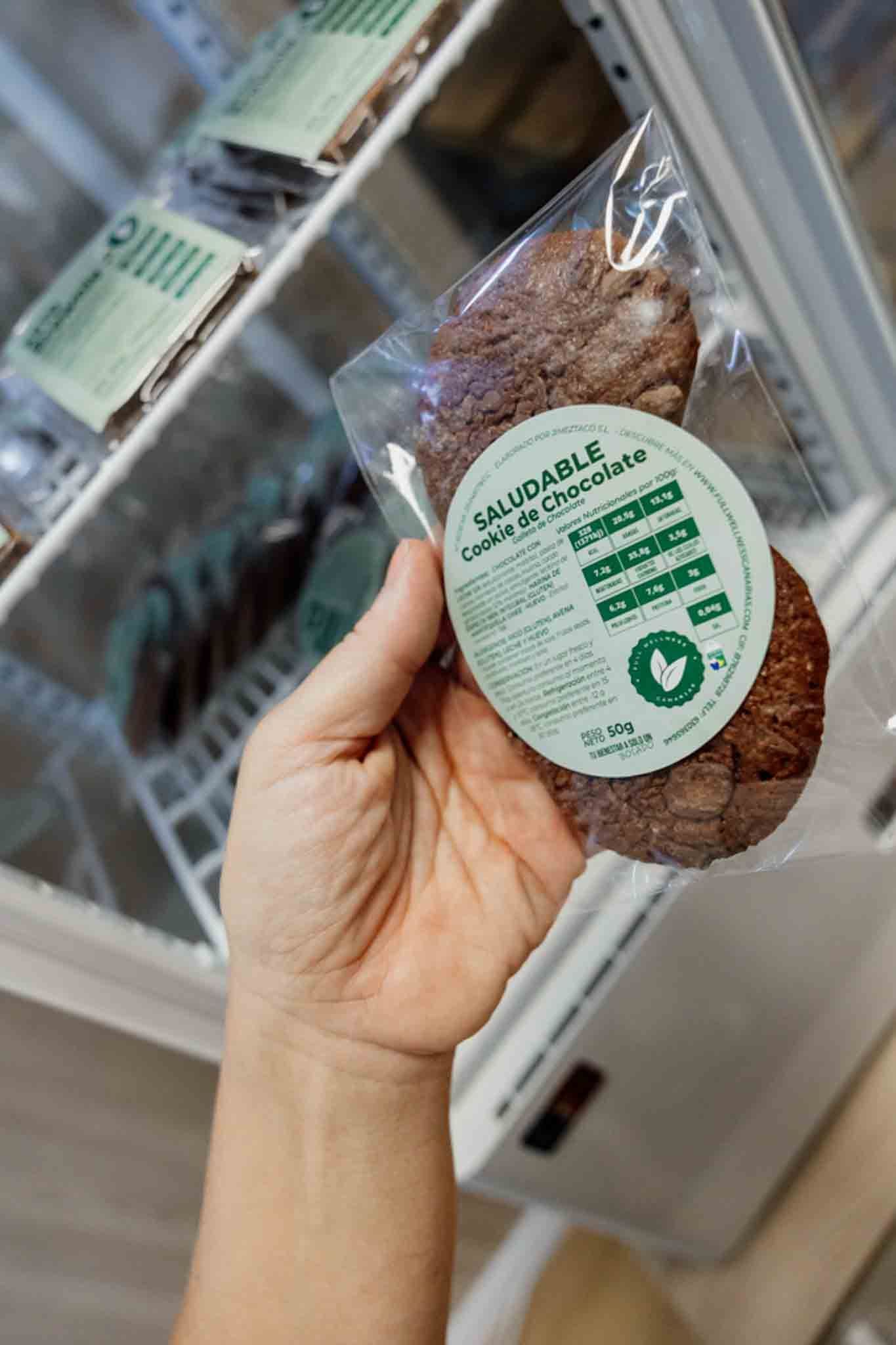 Una mano sostiene un paquete de dos galletas redondas de chocolate negro en el refrigerador. La etiqueta es verde y blanca.
