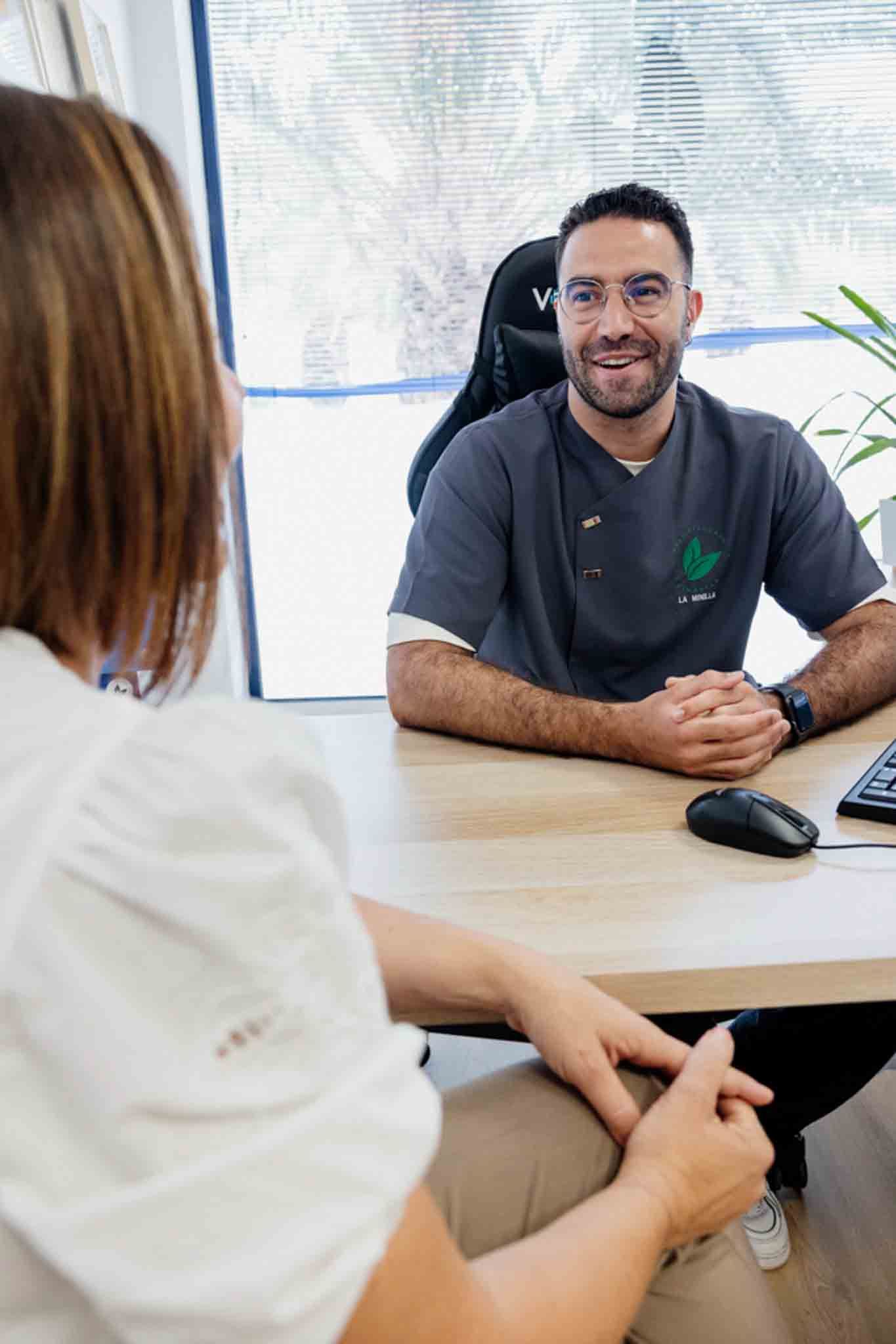 Un médico sonríe a un paciente en su consultorio. Lleva barba, gafas y una camisa gris con un logotipo.