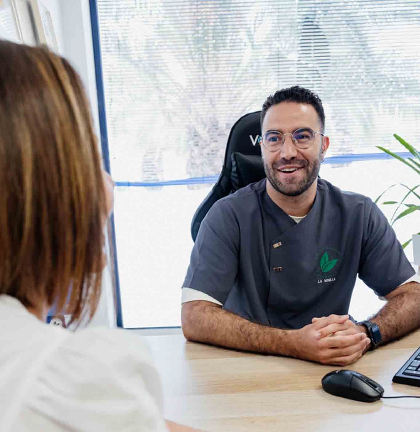 Un médico sonríe mientras observa a un paciente. Lleva gafas y un uniforme gris, sentado en un escritorio en una oficina.