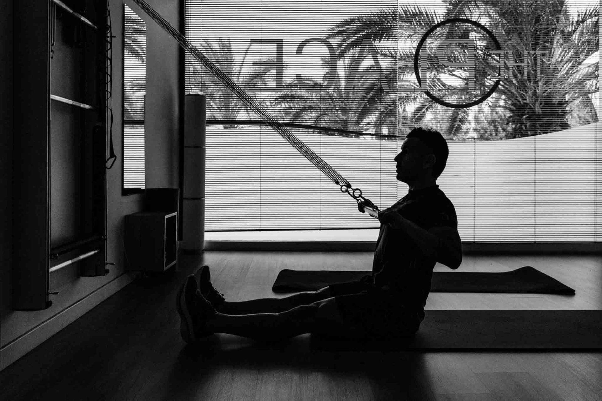 Silueta en blanco y negro de una persona sentada, tirando de un cable en una sala de ejercicios con una ventana al fondo.
