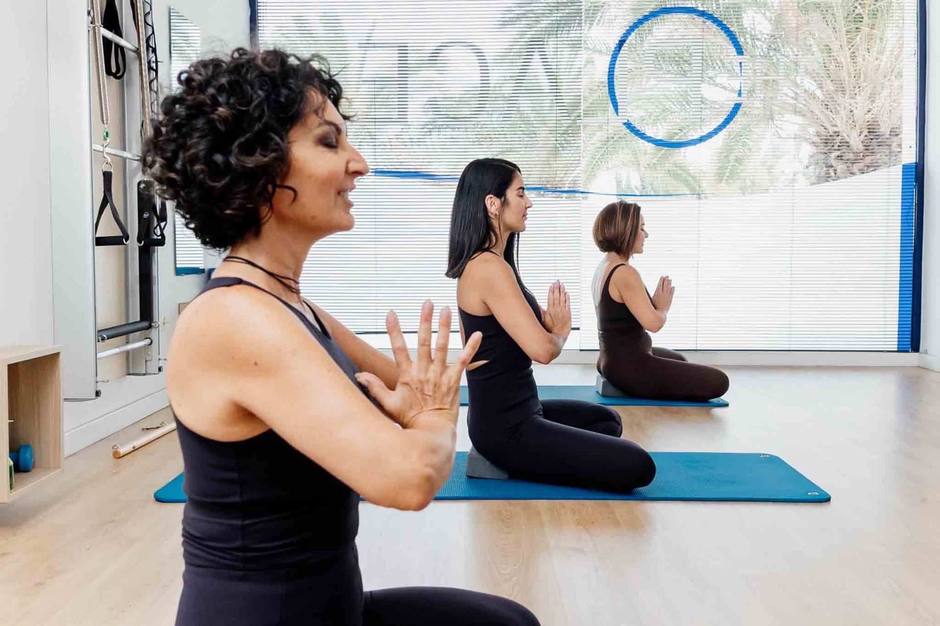 Tres mujeres en un estudio, meditando con las manos en oración. La luz del sol se filtra a través de una ventana tras ellas.