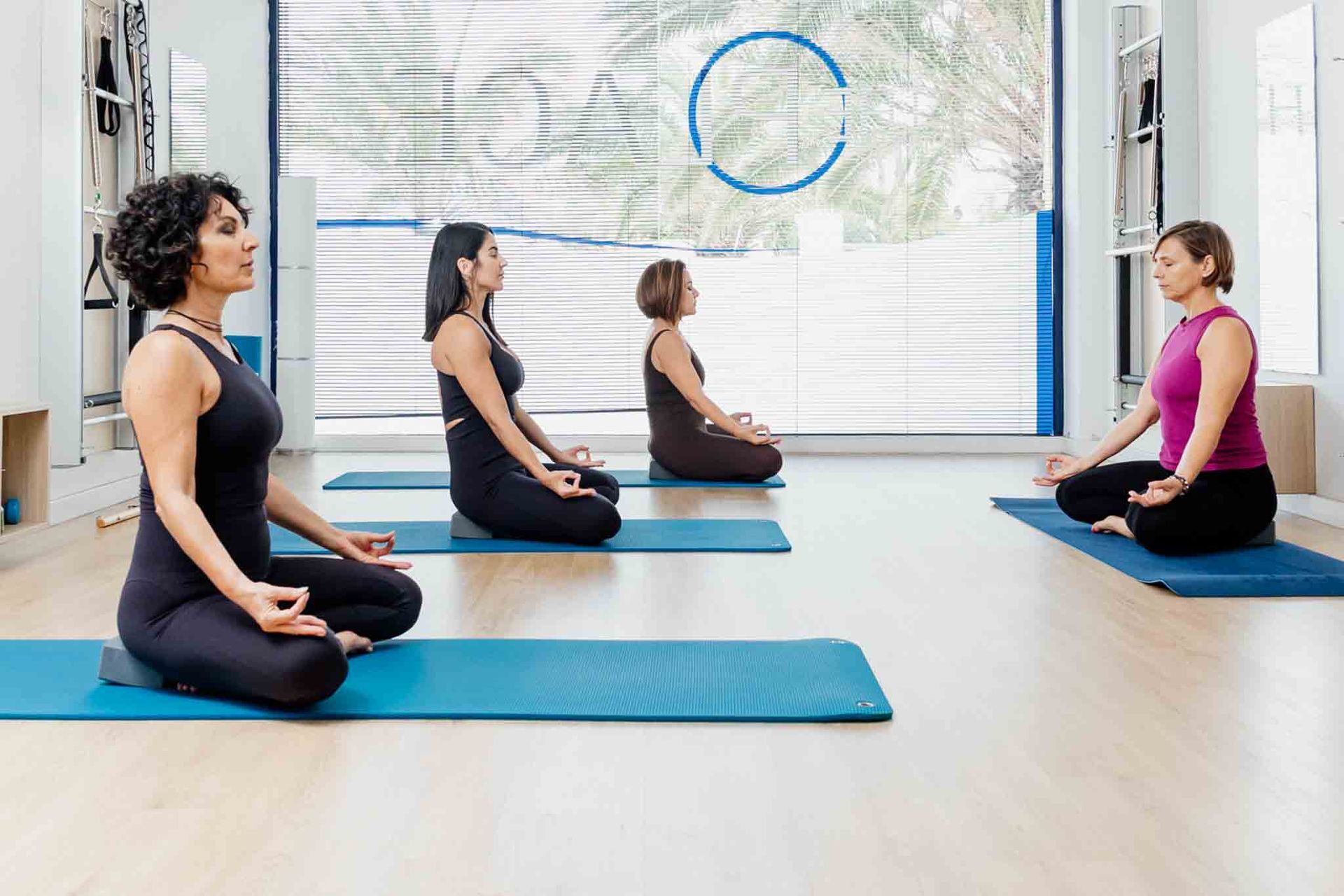 Cuatro mujeres meditan en una clase de yoga, sentadas con las piernas cruzadas sobre colchonetas azules. 