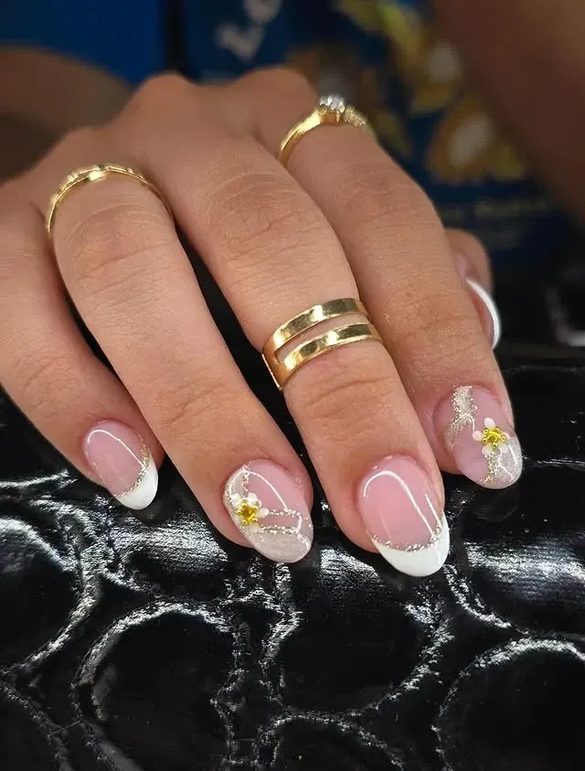 Mano cuidada con uñas de punta francesa, adornada con anillos dorados y acentos florales.