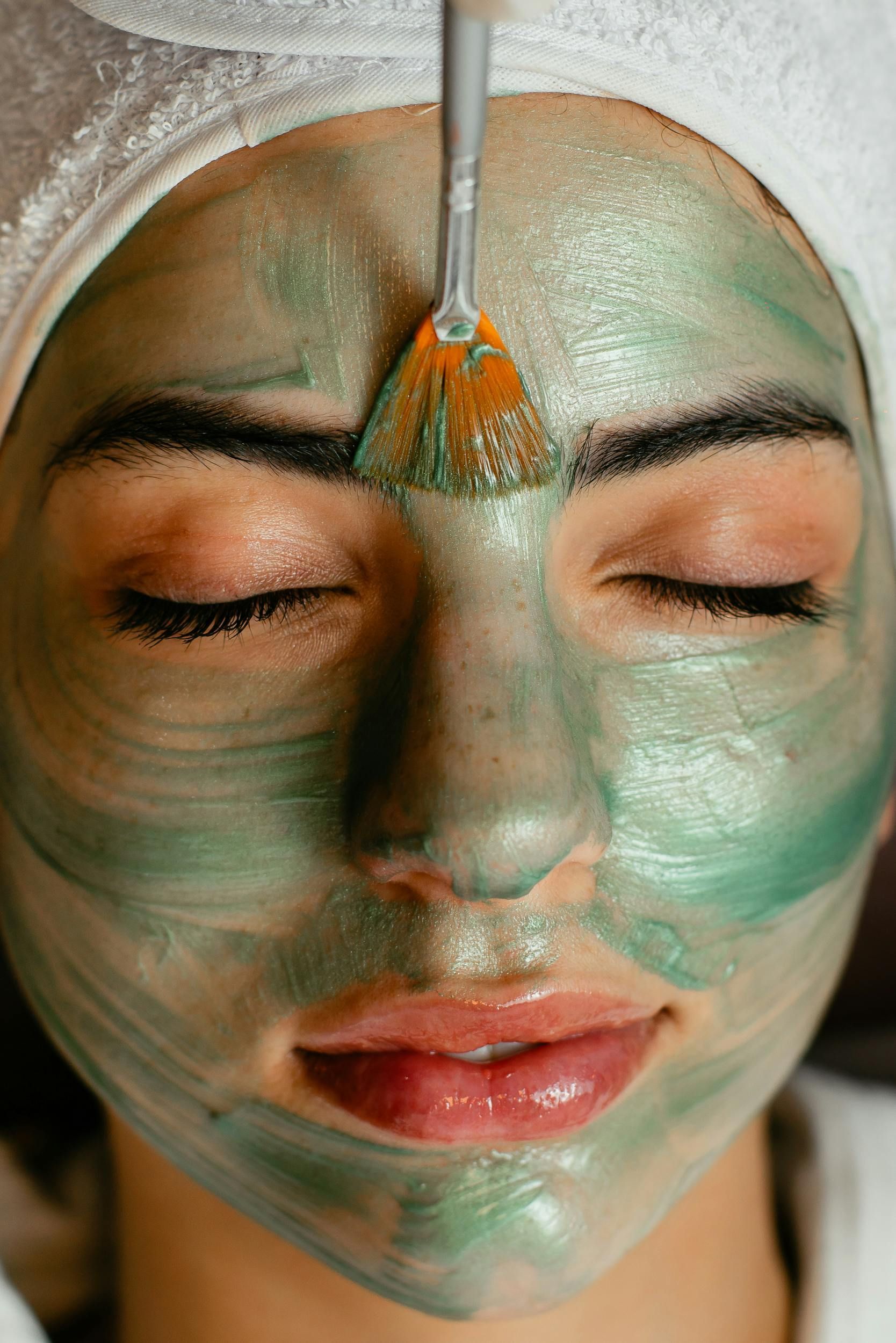 Mujer con los ojos cerrados, con mascarilla verde aplicada en el rostro, siendo cepillada con un aplicador cosmético.