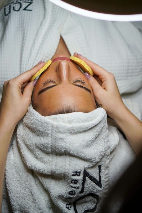 Mujer recibiendo un tratamiento facial en un entorno de spa. Sosteniendo esponjas sobre los ojos, vestida con bata y toalla.