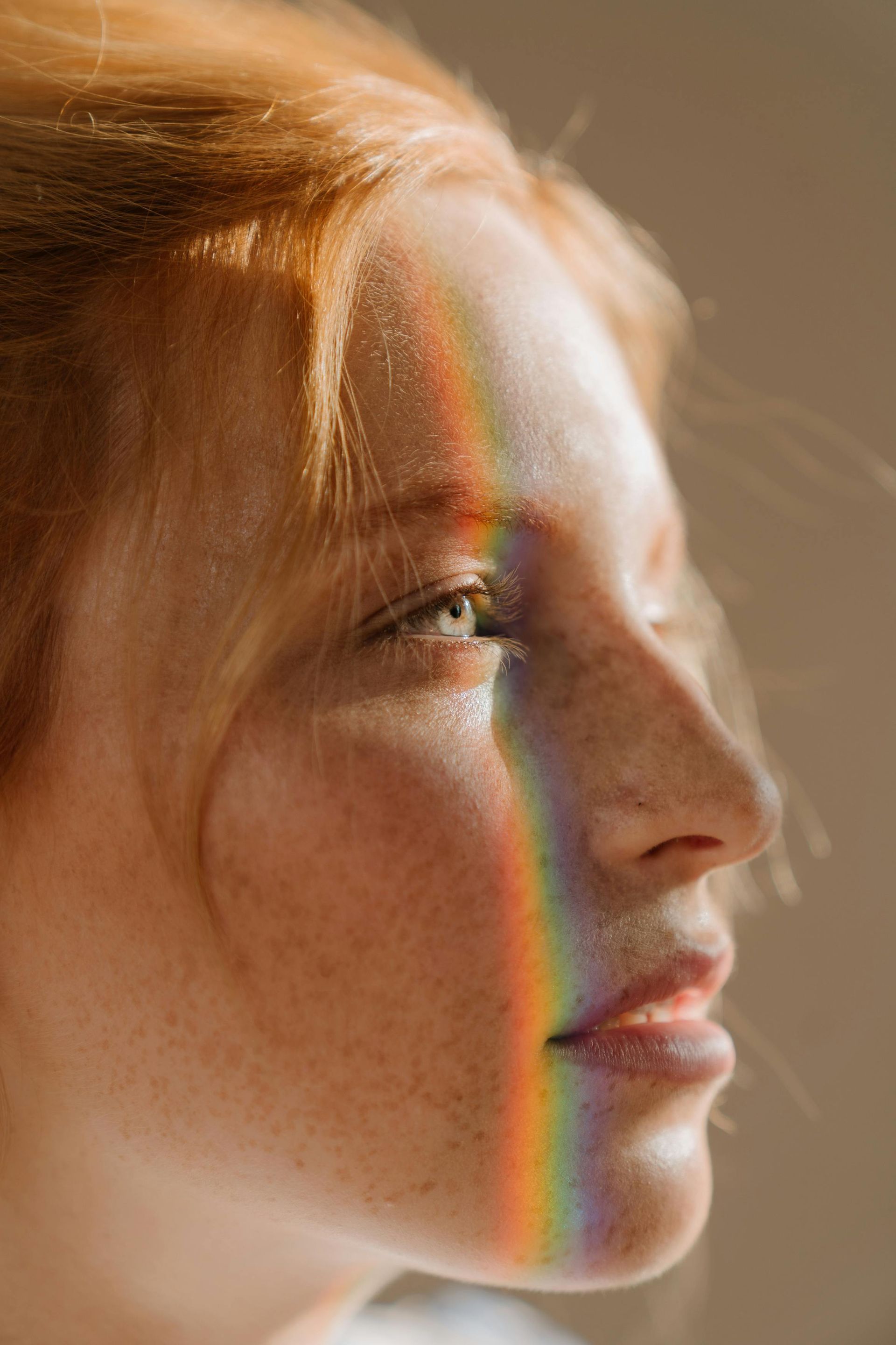 Mujer con cabello rojo, pecas y una luz de arco iris en su rostro, de perfil.