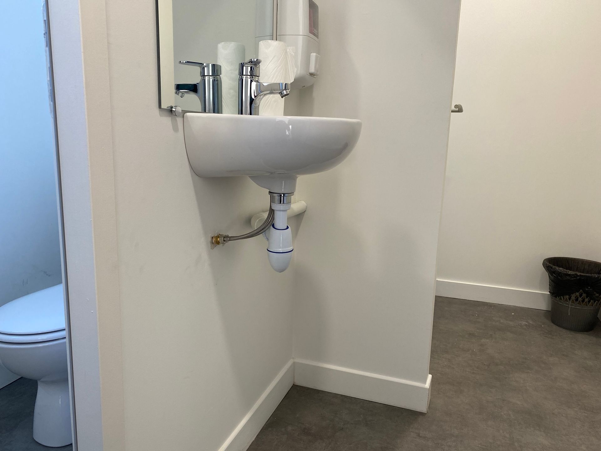 Des toilettes avec un lavabo, un miroir et une cuvette visibles. Les murs sont blancs et le sol gris.