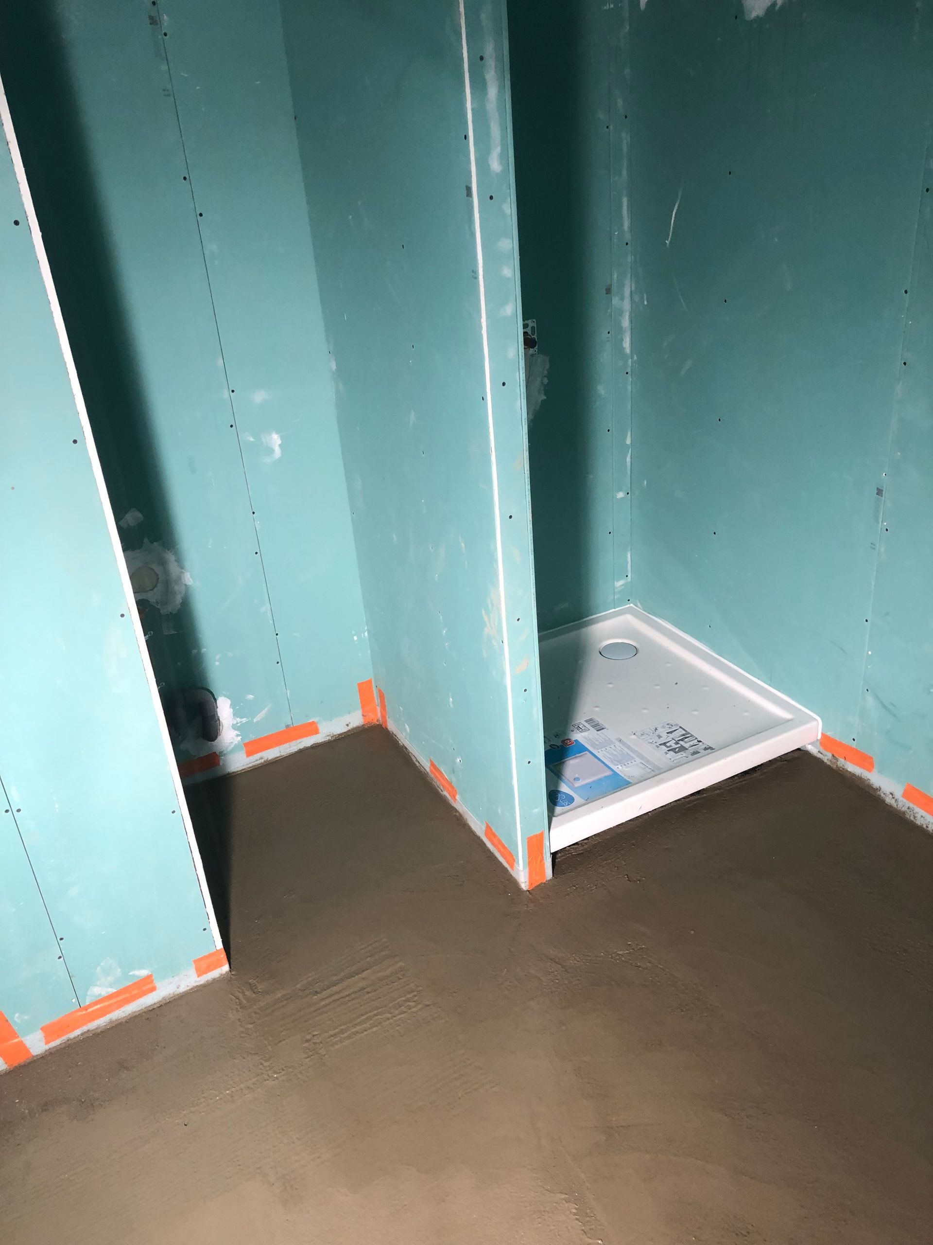 Intérieur d'une douche en construction ; murs en plaques de plâtre bleues, receveur de douche blanc, sol en ciment.