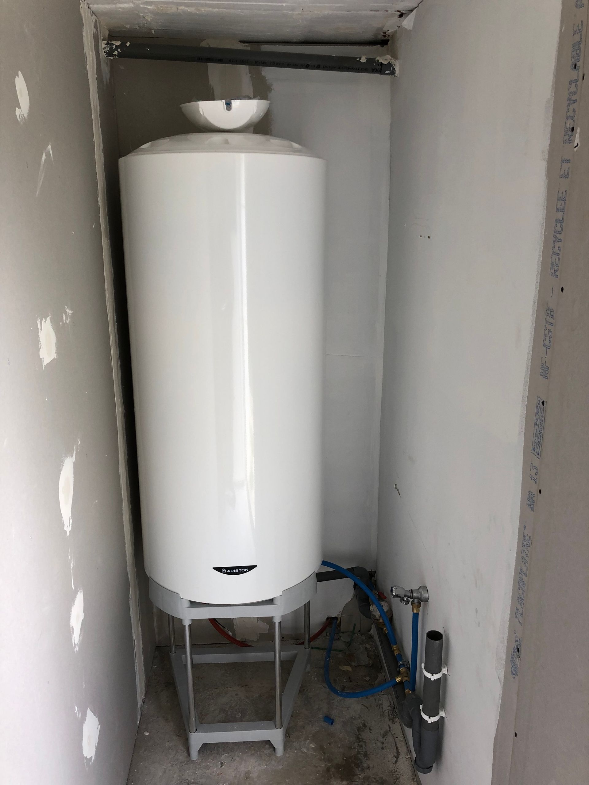 Chauffe-eau blanc sur un support dans une petite pièce inachevée, avec des tuyaux et des fils électriques apparents.