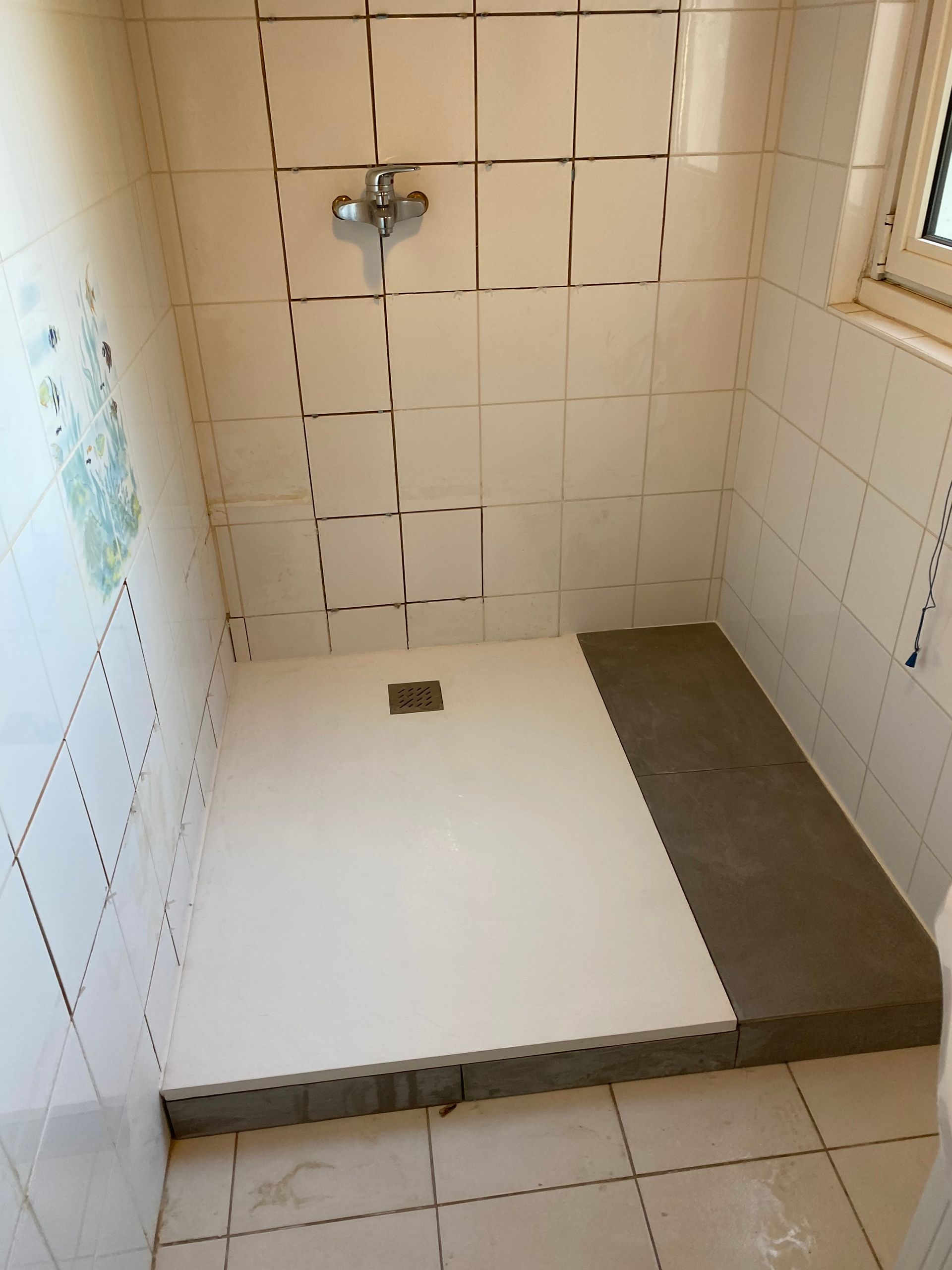 Douche carrelée de blanc avec un receveur rectangulaire blanc et gris et un robinet argenté.