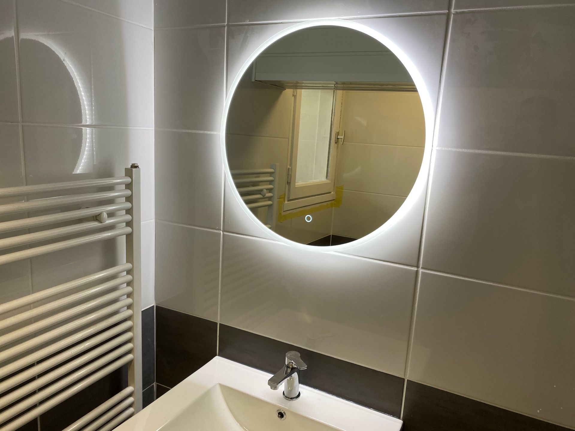 Salle de bains avec miroir rond à éclairage LED, lavabo et sèche-serviettes. Murs carrelés en noir et blanc.
