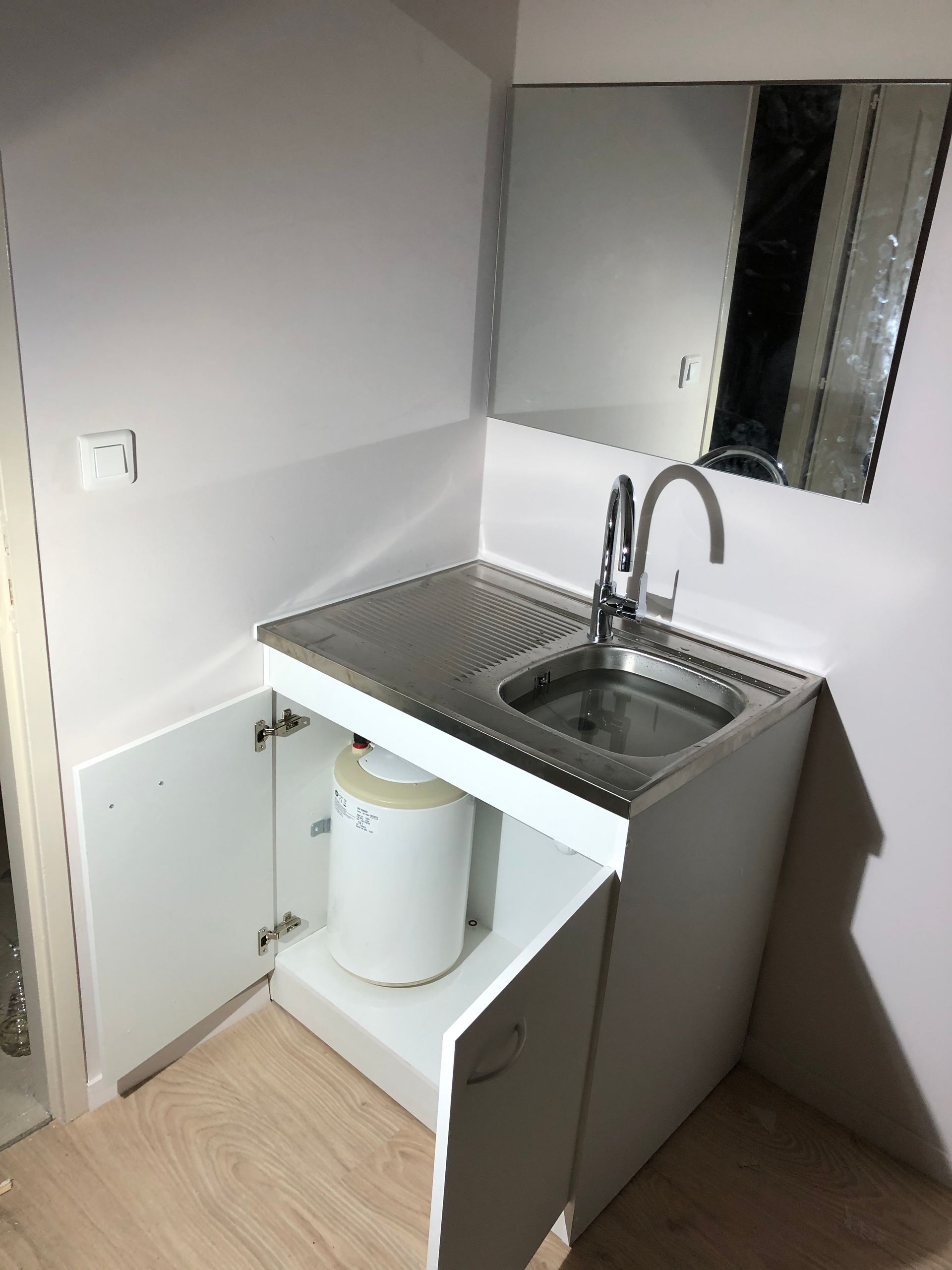Petit meuble sous-évier blanc avec évier en acier inoxydable, miroir et porte ouverte révélant un chauffe-eau blanc.