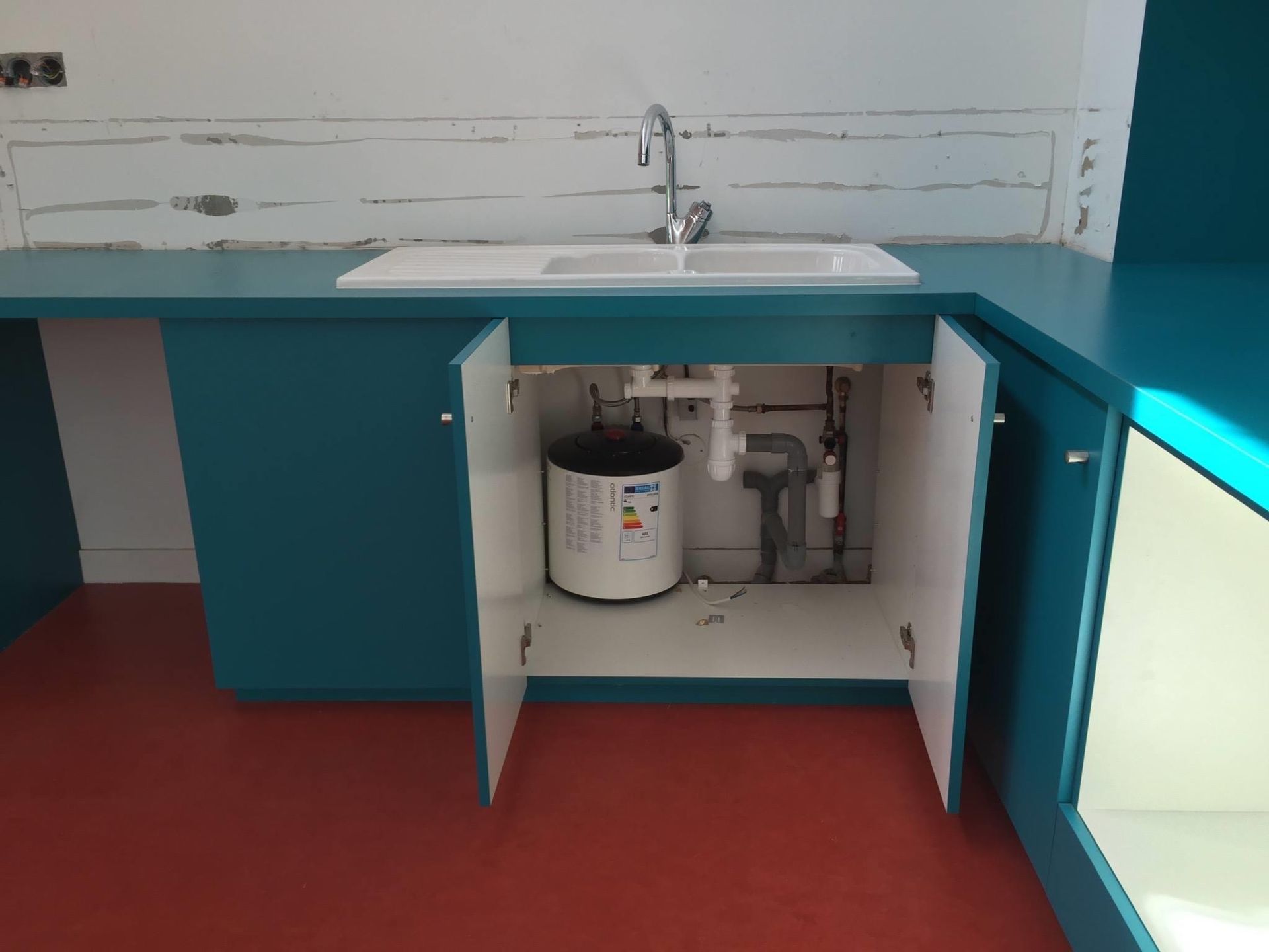 Meuble bleu à portes ouvertes sous un évier blanc, laissant apparaître la plomberie et un chauffe-eau, sol rouge.