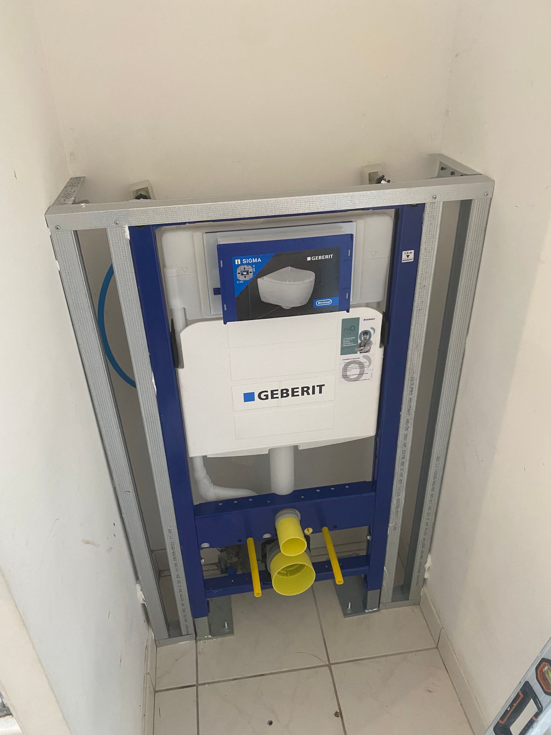 Dans une petite salle de bains, une structure métallique soutient un réservoir de toilettes bleu et blanc. On aperçoit des tuyaux jaunes.