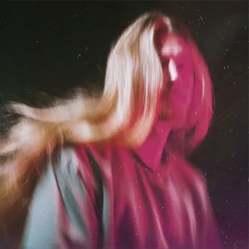 Figura borrosa con cabello largo rubio y rosa sobre un fondo oscuro, en movimiento.