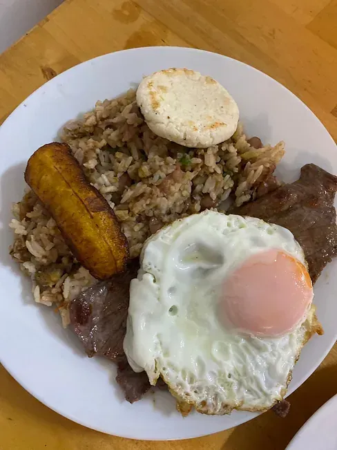 Un plato de comida con un huevo frito, arroz, carne y un plátano en una mesa de madera.