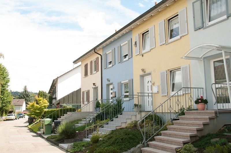 Eine Reihe pastellfarbener Stadthäuser mit Eingangstreppen und kleinen Gärten an einer sonnigen Straße