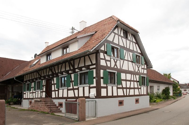 Traditionelles Fachwerkhaus mit weißen Wänden, dunklen Holzbalken, grünen Fensterläden und einem roten Ziegeldach an einer Straße.