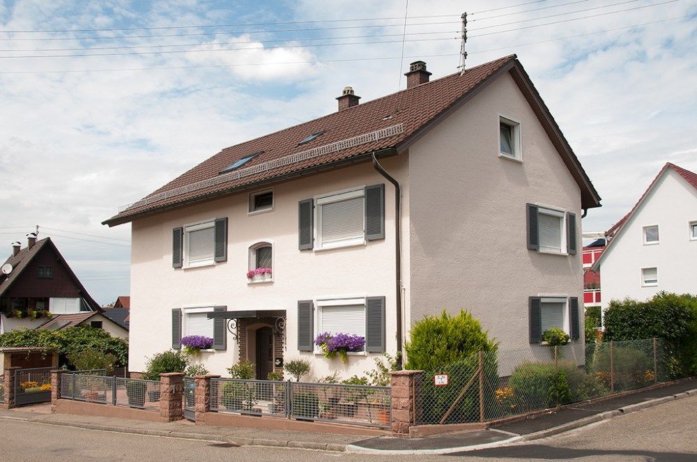 Zweistöckiges cremefarbenes Haus mit rotem Dach und lila Blumen davor, auf einem Eckgrundstück in einem Vorort