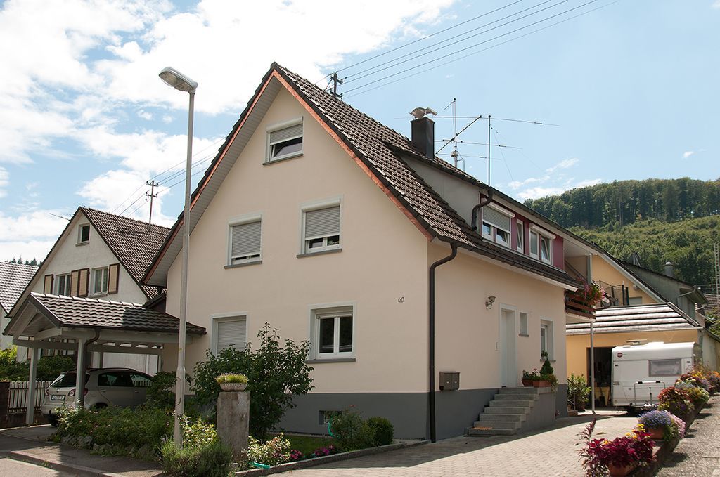 Ein cremefarbenes Haus mit rotem Ziegeldach in einem sonnigen Wohngebiet, mit Blumen und einem Carport.
