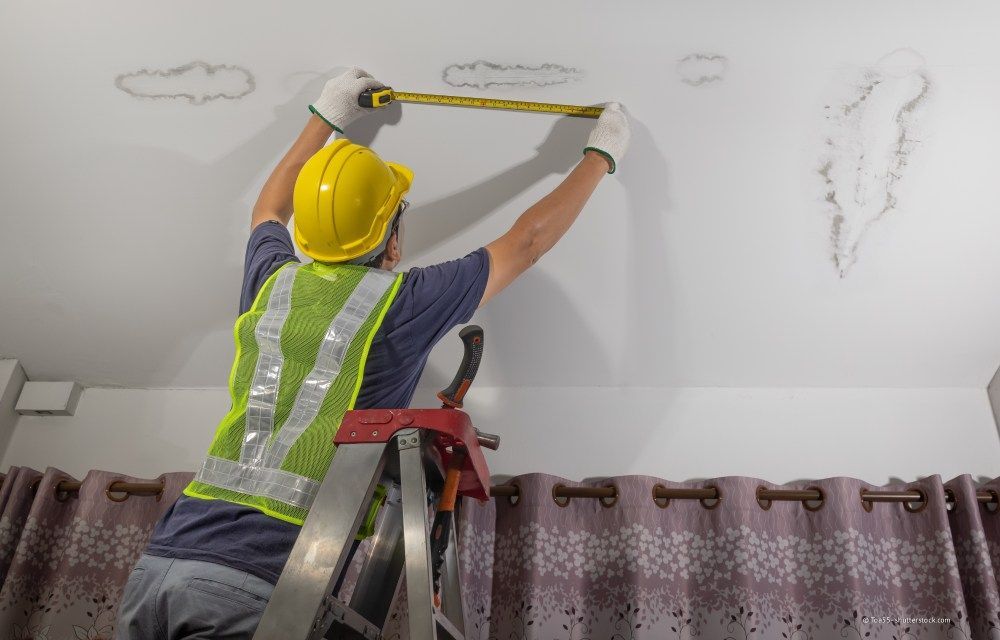 Ein Arbeiter auf einer Leiter misst mit einem Maßband die Wandhöhe nahe der Decke; er trägt einen Schutzhelm und eine Sicherheitsweste.