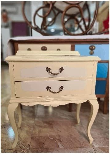 Una pequeña mesita de noche blanca con dos cajones está sobre un suelo de baldosas.