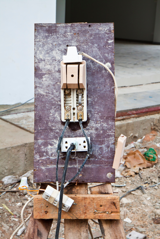 Panneau électrique de fortune sur une planche de bois avec des fils exposés, un disjoncteur et une prise, à l'extérieur.