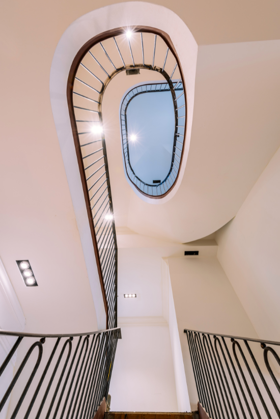 Escalier en colimaçon avec rampe sombre, murs blancs et éclairage zénithal.