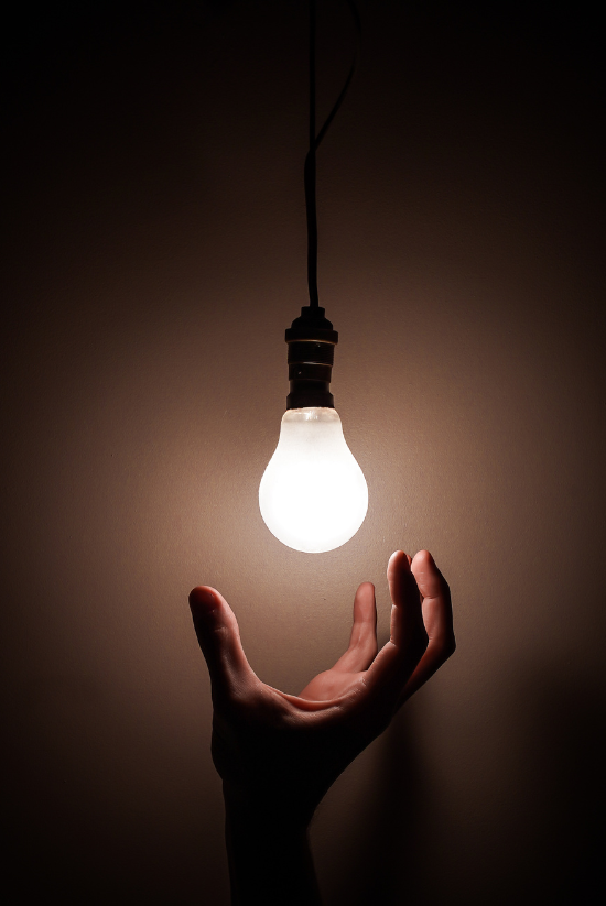 Main tendue vers une ampoule lumineuse suspendue à un cordon, dans une pièce sombre.