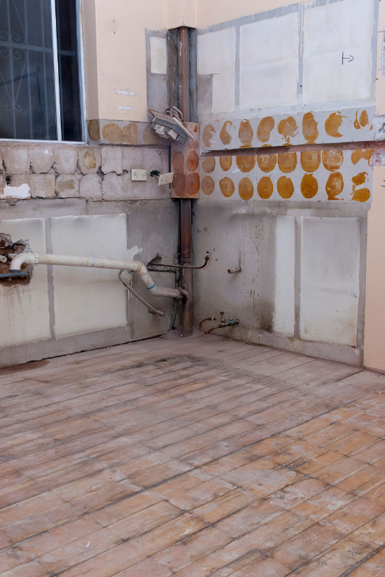 Rénovation de cuisine : murs, tuyauterie et parquet apparents. Restes de cheminée, de fenêtre et de carrelage bruns visibles.