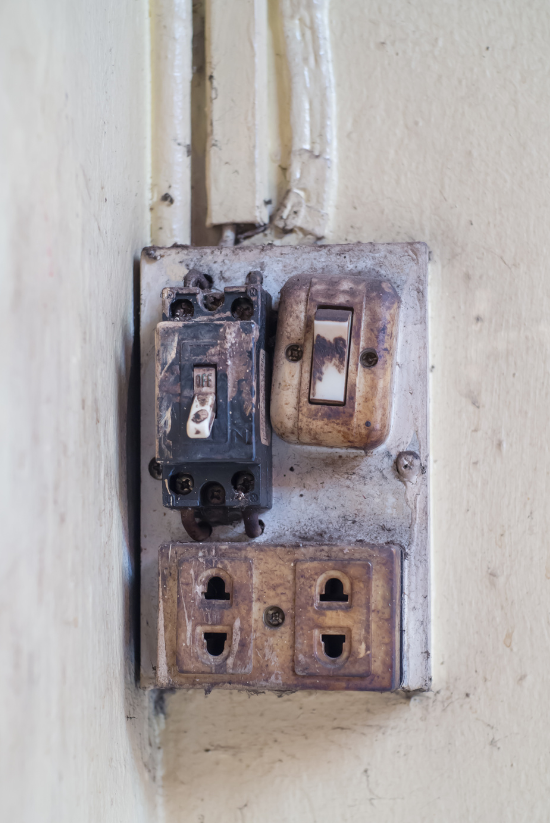 Vieille prise électrique, interrupteur et disjoncteur usés par les intempéries sur un mur beige.