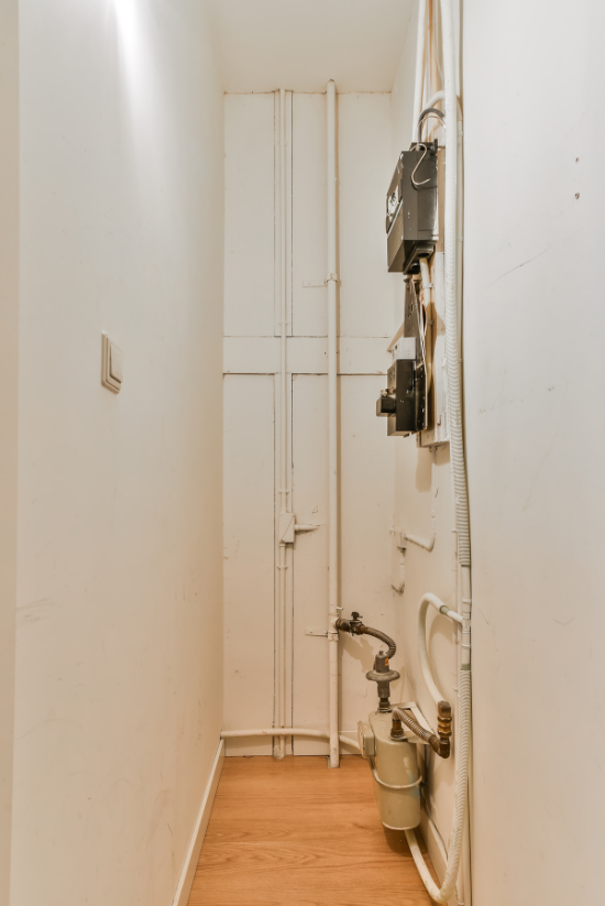 Buanderie étroite avec compteur de gaz, boîtes électriques, tuyaux et plancher de bois.