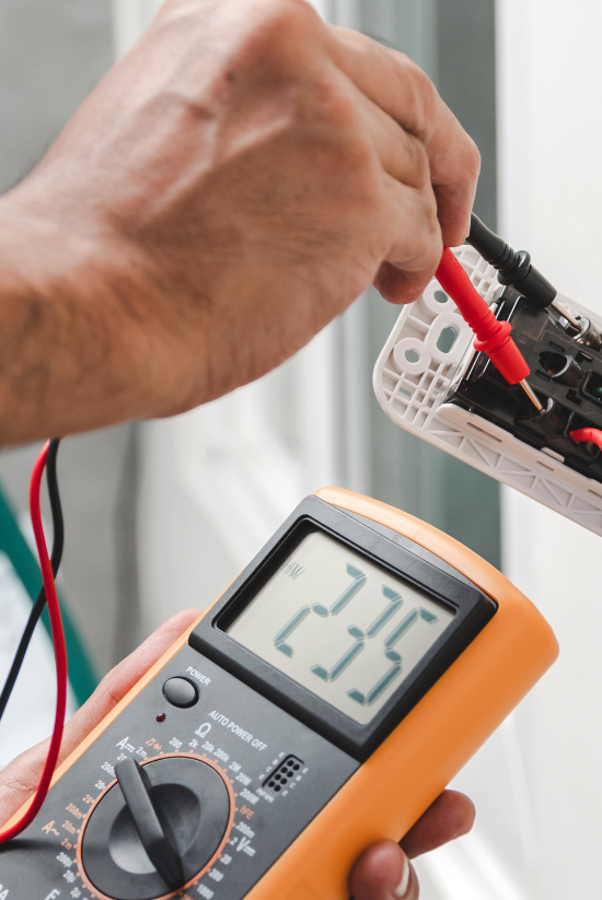 Personne testant la tension d'une prise électrique avec un multimètre, l'écran affiche 235.