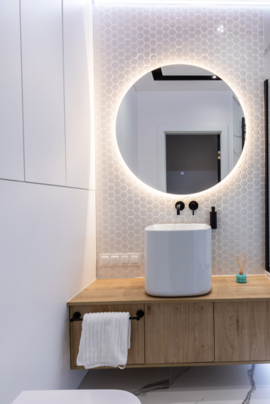 Salle de bain moderne avec un miroir rond rétroéclairé, un lavabo blanc et une vanité en bois.