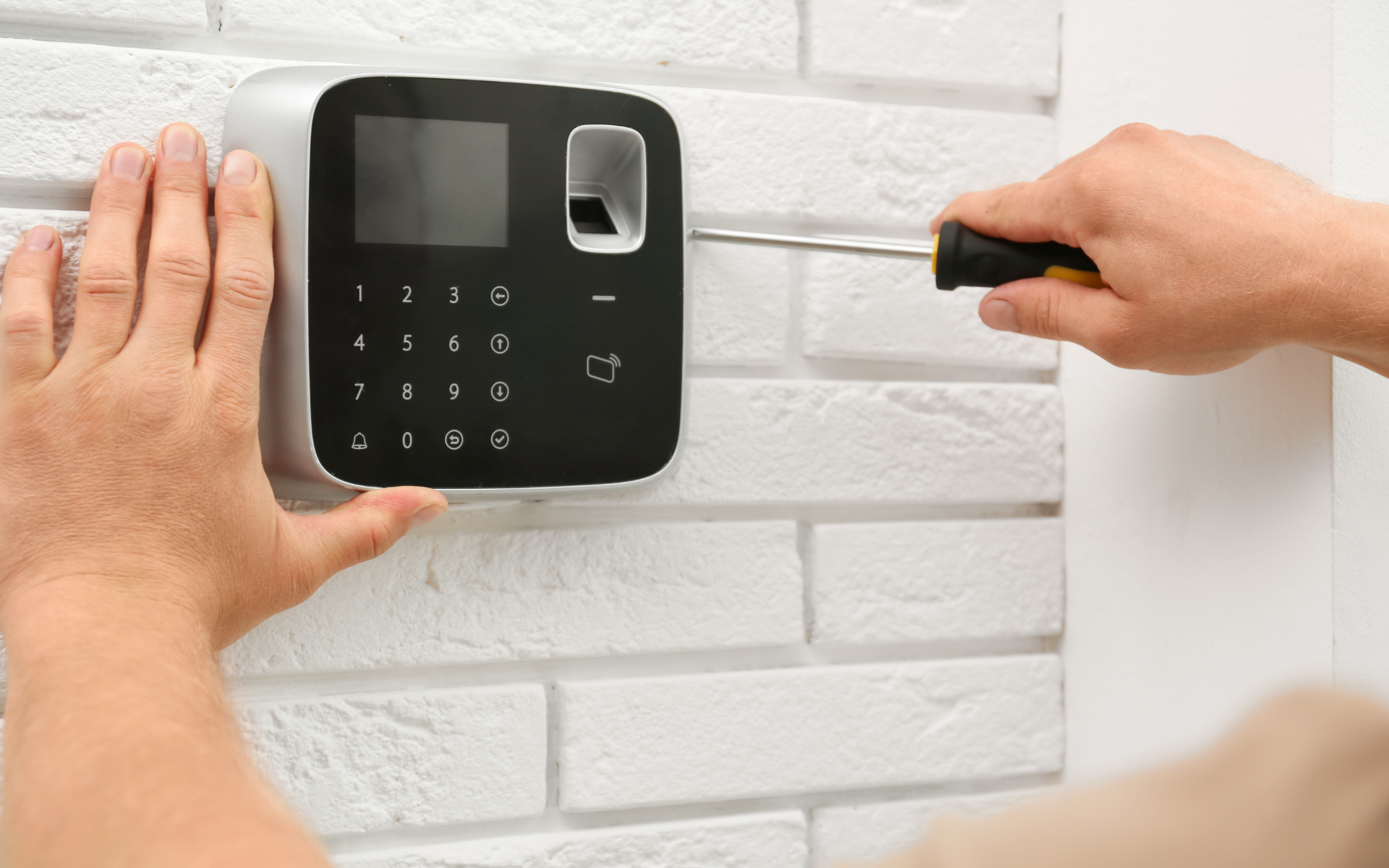 Personne installant un scanner d'empreintes digitales avec un tournevis sur un mur de briques blanches.