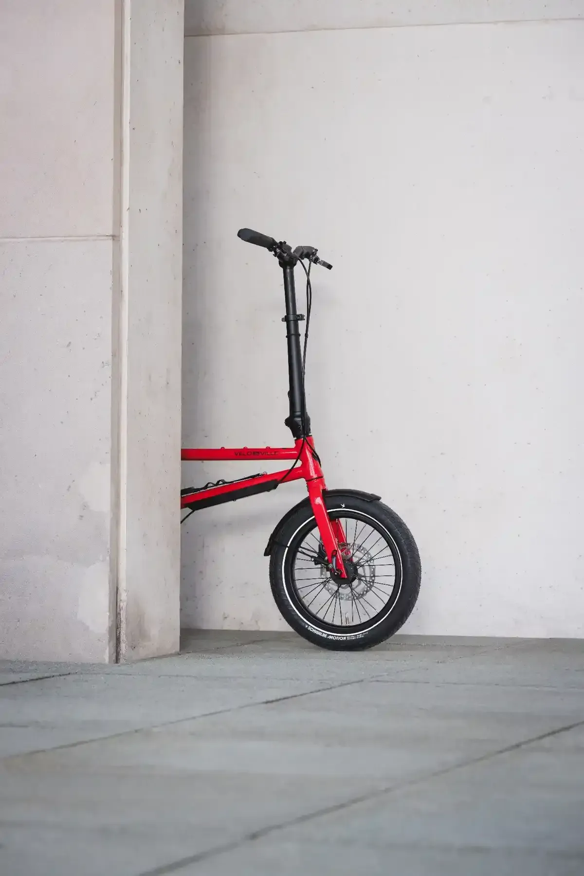 Un patinete eléctrico plegable rojo aparcado contra una pared blanca en una acera gris.
