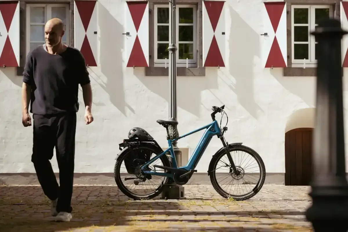 Una persona pasa caminando junto a una bicicleta azul frente a un edificio con contraventanas rojas y blancas.